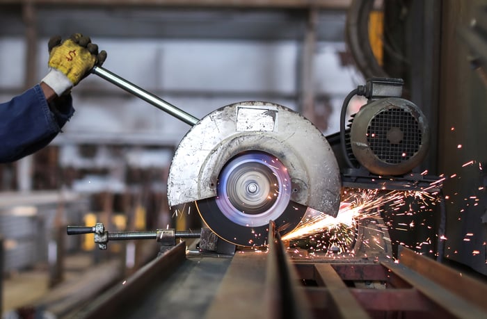 A person with a glove that has a dollar sign on it grips a circular saw in a machine shop.
