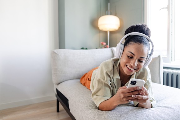 Happy person using headphones and a phone while lying on a couch.