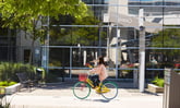 Lady biking on Google campus with Google name reflected on window in background. IS GOOG.