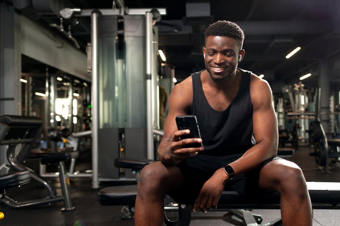 Someone at a gym is seated, looking at a phone and smiling.