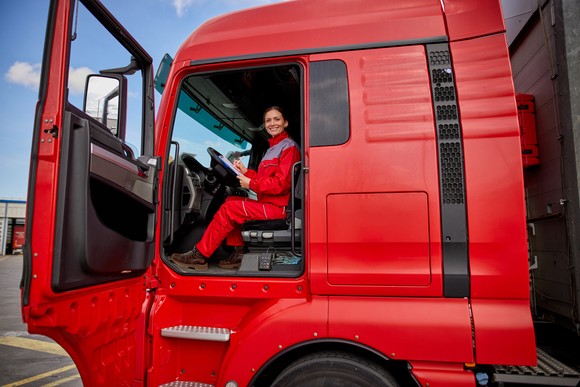 Person in red uniform sitting at the wheel of a big red tractor trailer semi truck.