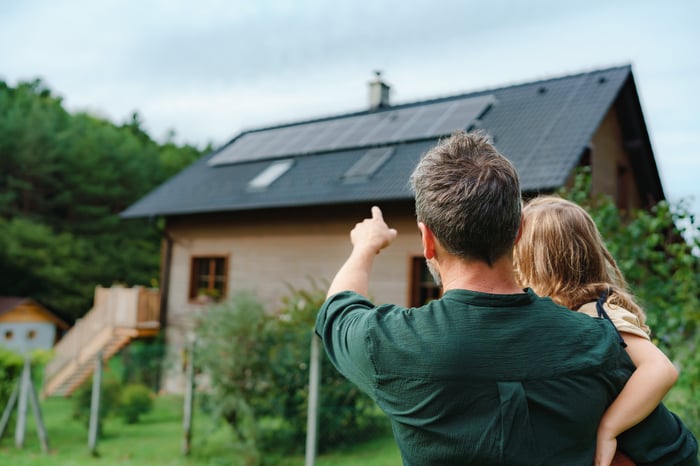 Adult with child in arms pointing to a house with solar panels on its roof.