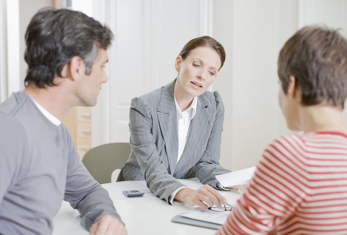 A person points to papers in a binder while sitting at a table with two other people.