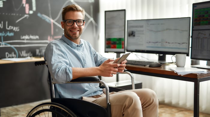 An investor sits in front of a desk with multiple computer monitors, holding a tablet.