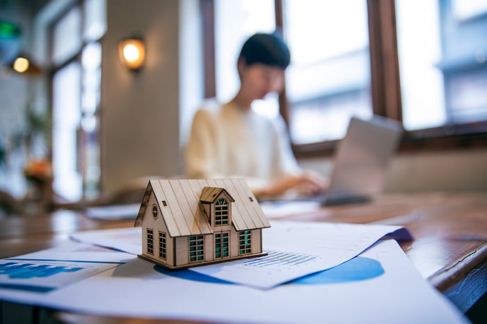 Small model home on a desk, with person working on laptop in the background.