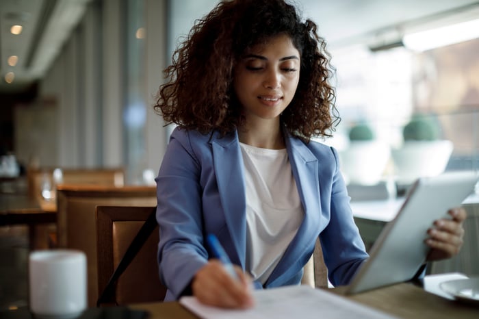 An investor sitting at a table and holding a tablet while writing on a piece of paper.