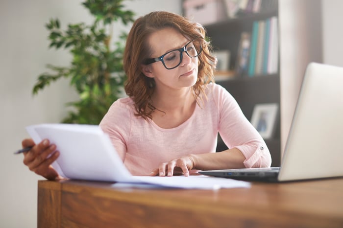 An investor holds a sheaf of papers while looking at a laptop and sitting at a desk.