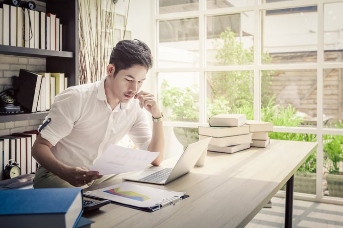 An investor sitting in an office in front of a laptop ponders some data that's printed out on a piece of paper.