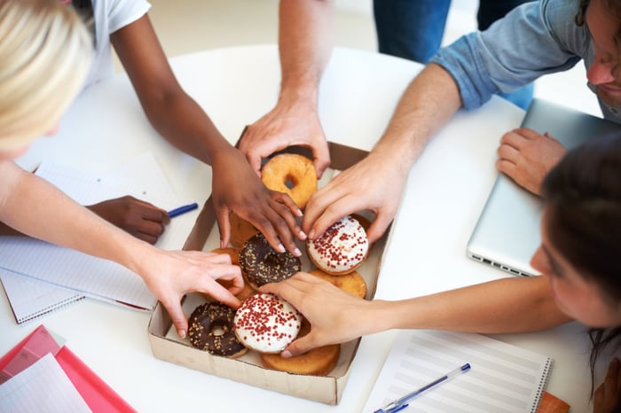 People around a table, taking doughnuts from a box.