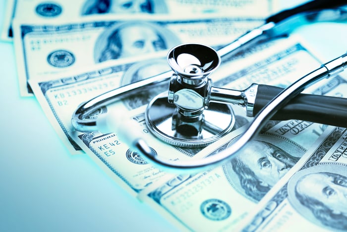 A stethoscope sitting atop hundred-dollar bills spread out on a table.