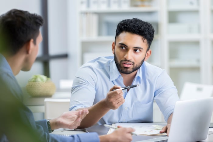 Two investors sit at a desk in front of laptops and papers as one holds a pen while explaining something to the other.