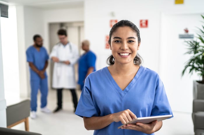 A healthcare professional holds a tablet while smiling in a doctor's office.