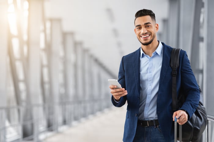 Someone is walking through an airport with a suitcase, smiling and holding a phone.