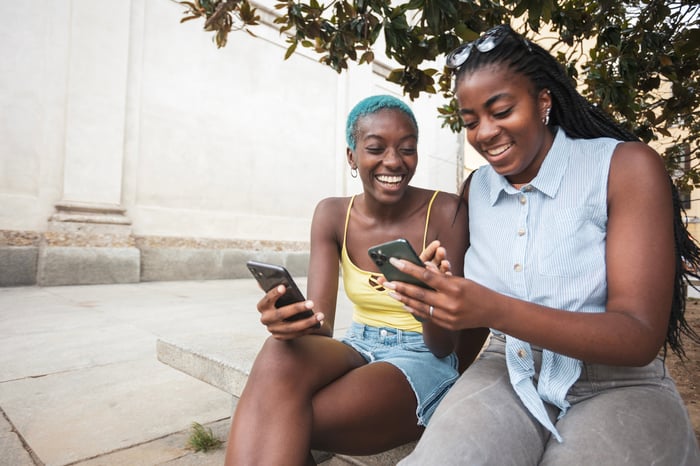 Two friends smiling while checking their smartphones.
