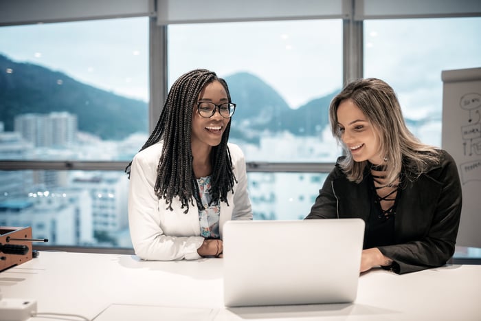 Two people smile in an office with a Brazilian landscape visible from the window.