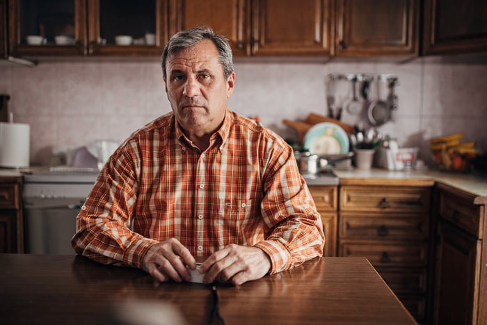 A frowning person in a plaid shirt sitting at a kitchen table.