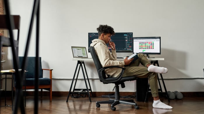 A young person taking some down time and reading a book beside a desk with two active computer screens.