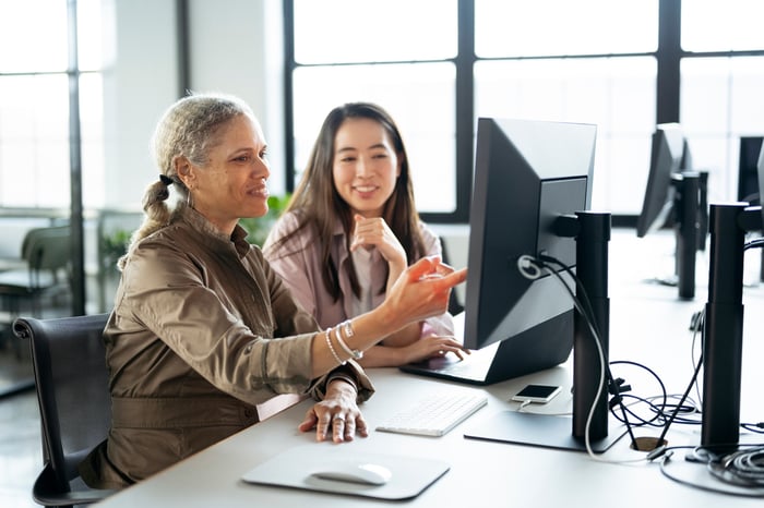 Two professionals smiling and talking while one points to a desktop on a table.
