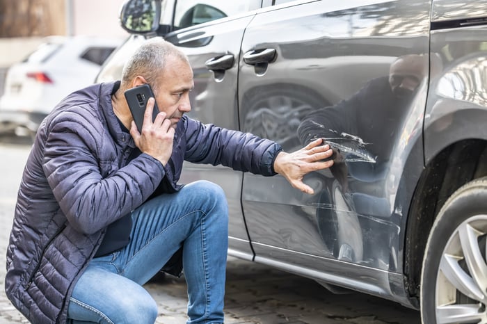 A person looks at a dent in their car while talking on the phone.