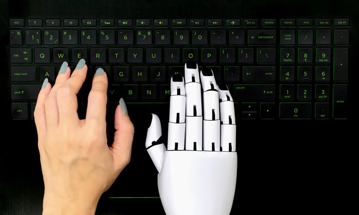 A human hand and a robot hand resting side by side on a black computer keyboard.