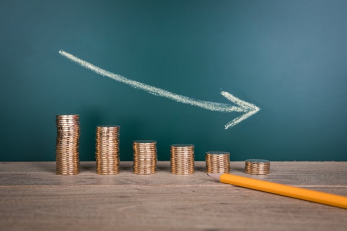 A descending stack of coins with an arrow pointing down drawn above them and a pencil in the foreground.