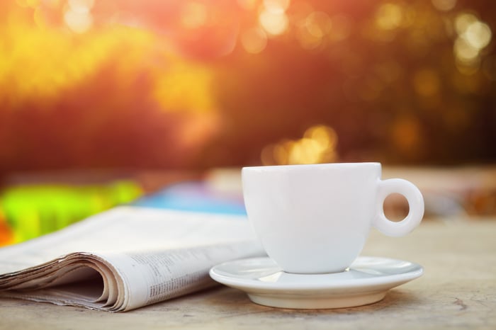 A coffee cup sits beside a newspaper on an outdoor table with dappled sunlight in the background.
