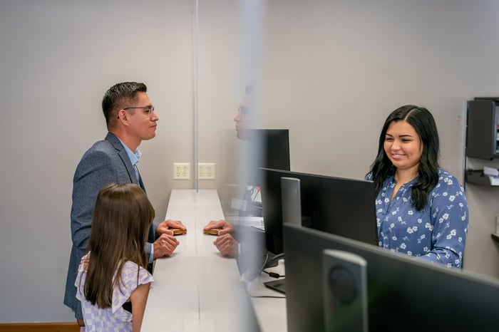 Man and his daughter stand being helped by a bank teller. 