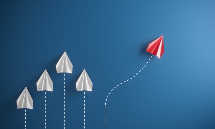 A red paper airplane going in a different direction than a group of other paper airplanes, illustrating the concept of contrarian investing in the stock market.