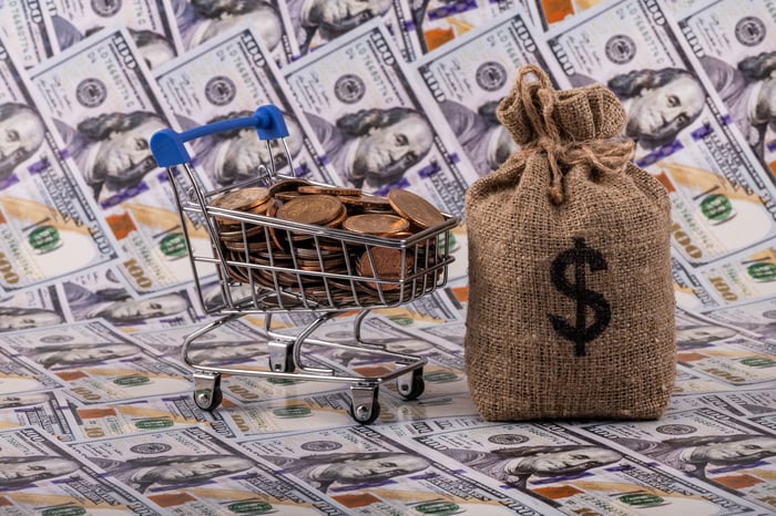 A shopping cart full of pennies next to a bag of money on $100 bills.