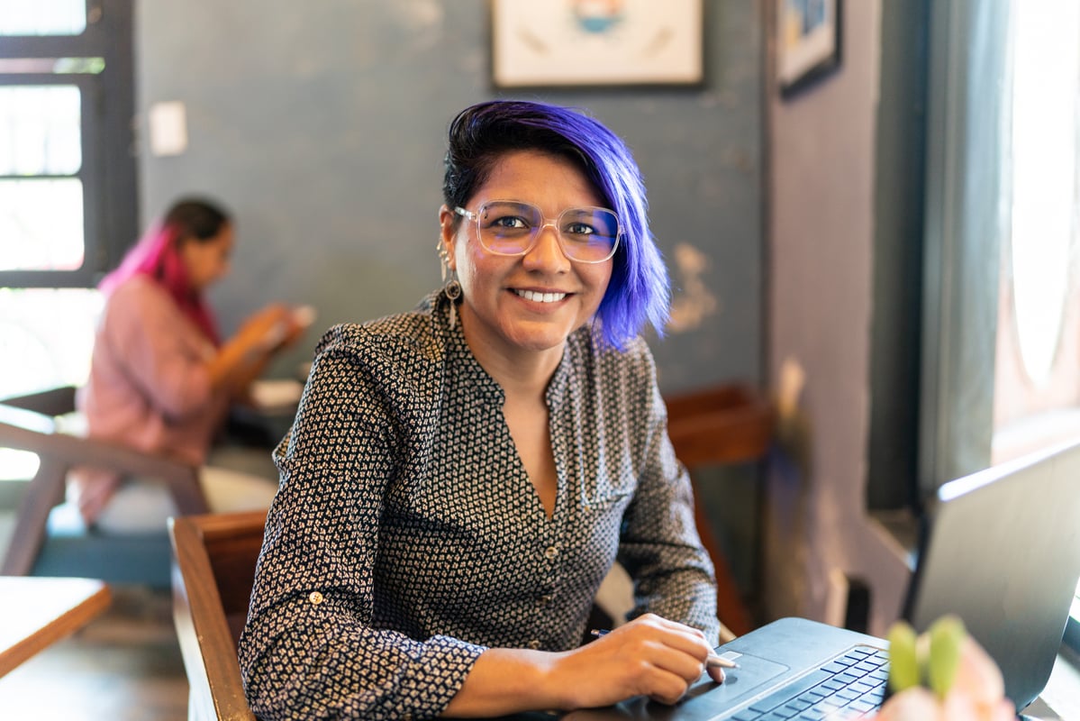 Businesswoman_with_vibrant_purple_hair_and_glasses_smiles_while_working_on_a_laptop_in_a_cafe_setting