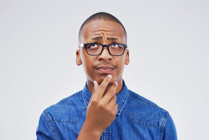 A person wearing glasses is looking up with a reflective look on their face and their hand on their chin.