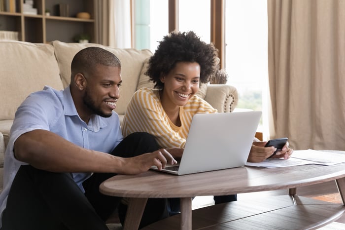 Two people smile as they look at a laptop in a living room.