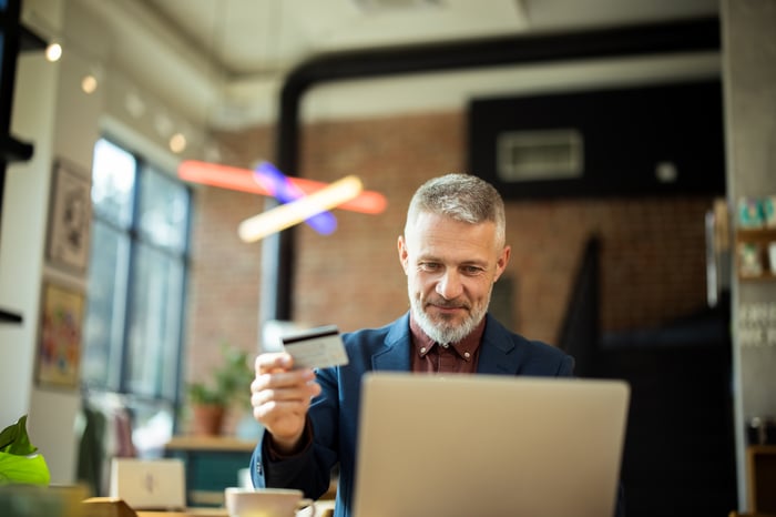 A person is sitting at a laptop and holding a credit card as they online shop in a cafe.