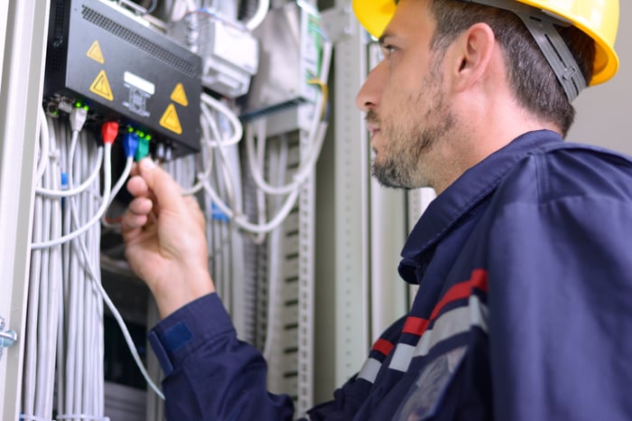 Electrician works a power box with wires coming out of it in a hardhat. 