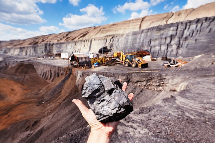 worker holds mineral in foreground with open pit mine in background. 