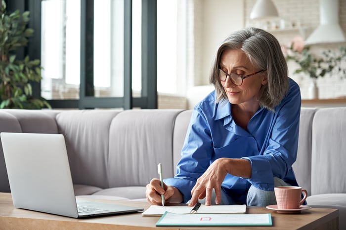 Person writing a note in notebook while glancing at laptop.