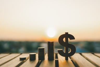 A U.S. dollar sign next to increasingly taller stacks of coins on a wood dock, illustrating the concept of growing passive income from investing in the stock market.