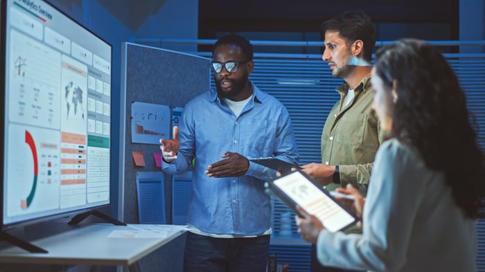 Three coworkers stand in front of a large computer screen. They are discussing the reports on the screen.