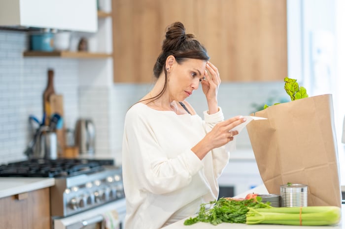 Person looking at a grocery list next to a pile of groceries in the kitchen. 