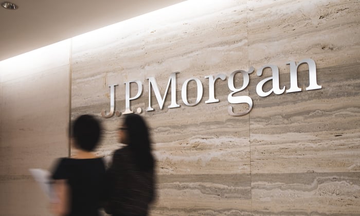 Inside JPMorgan office with JPMorgan logo on wall while two people walk by.