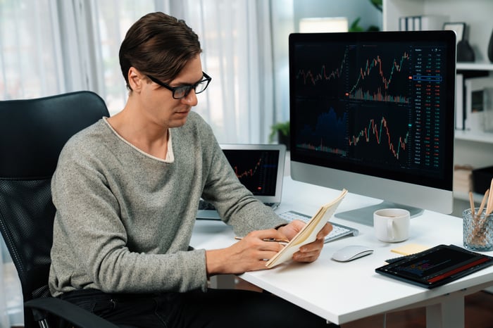 A person reviewing notes on a pad with graphs on a computer monitor in the background.