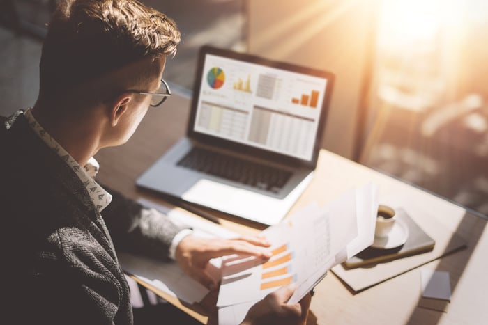 A person holds paper documents while looking at a computer.