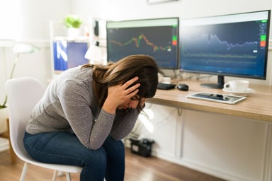 Person seated at a desk with two PC monitors holding head in hands.