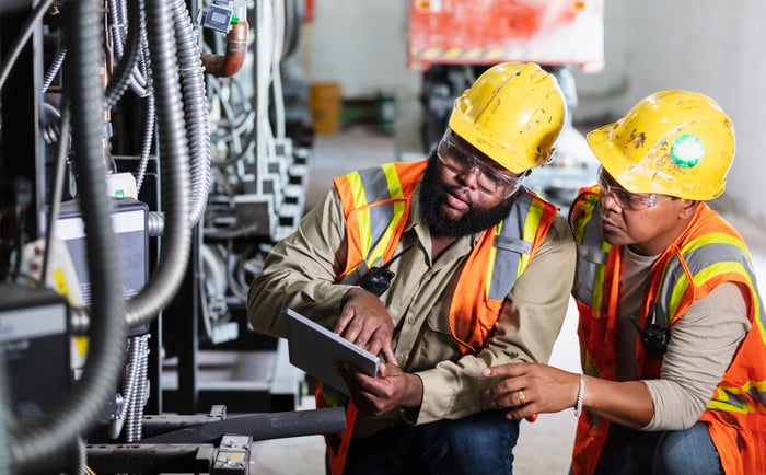 Two industrial workers look at an tablet next to hoses and wires.  