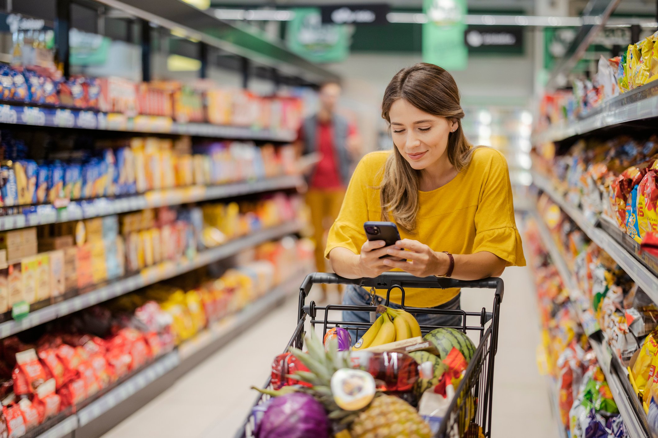 A person pushin a shopping cart down the grocery aisle while looking at a smartphone