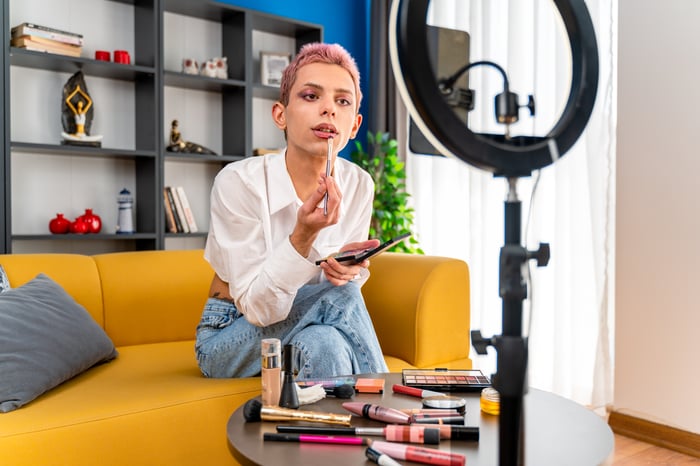 Woman sitting on couch in front of her phone and ring light putting on makeup. 