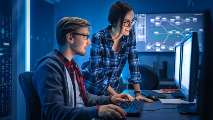 Two tech employees work in a dark data center in front of desktop computer.