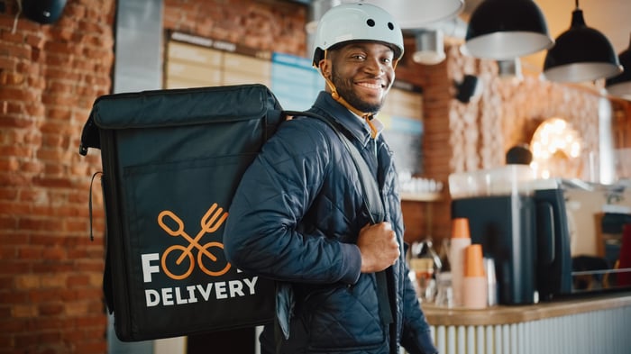 A delivery person has a backpack from a food delivery service on his back as he stands in a restaurant lobby