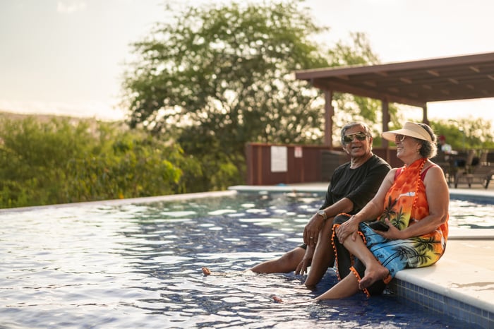 A couple on vacation sits poolside with their feet in the water.