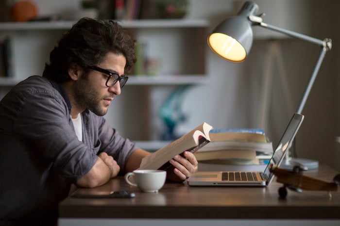 A student is reading a book near an open laptop. 